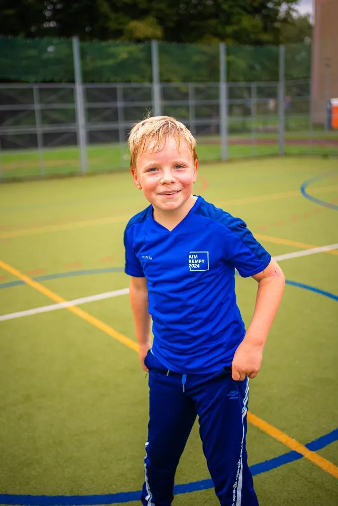 Skupina dětí v modrých tričkách drží společně trofej nad hlavami během venkovní sportovní akce.