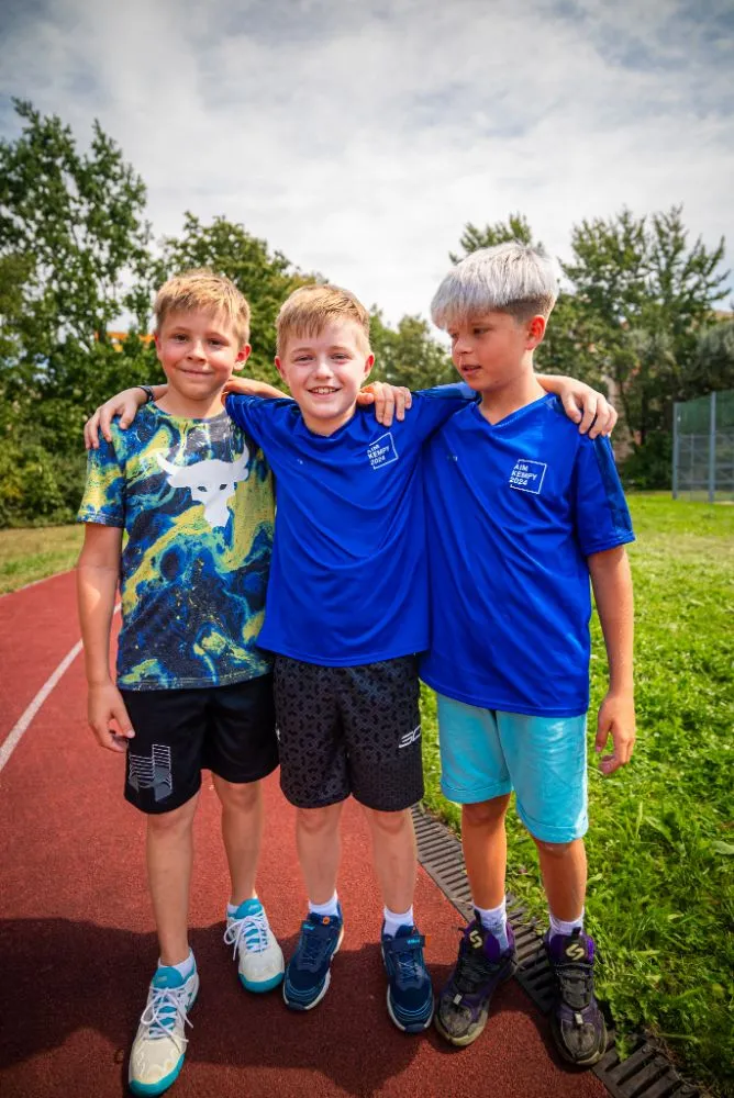 Skupina dětí v modrých tričkách drží společně trofej nad hlavami během venkovní sportovní akce.