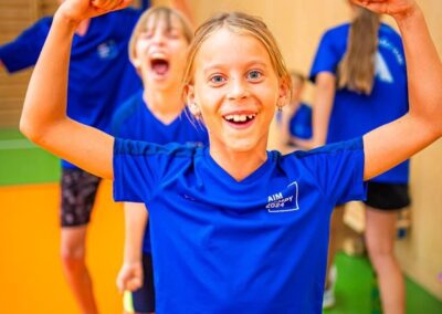 Dívka v modrém tričku ukazuje své svaly s nadšeným výrazem na obličeji, zatímco za ní další děti v tělocvičně oslavují.