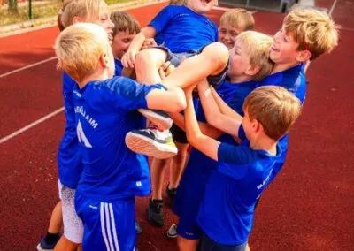Skupina chlapců v modrých tričkách na atletické dráze společně zvedá svého kamaráda do vzduchu.