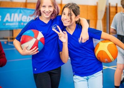 Dvě dívky v modrých tričkách, jedna drží basketbalový míč, druhá volejbalový míč, stojí v tělocvičně a usmívají se.