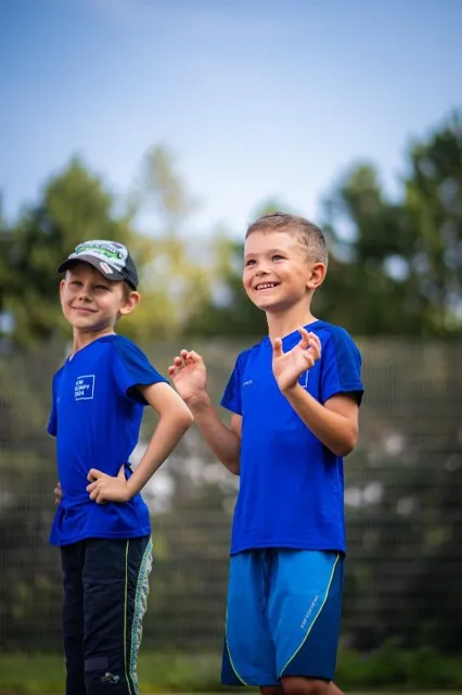 Dva chlapci v modrých tričkách stojí venku a smějí se během sportovní akce.