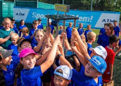 Skupina dětí v modrých tričkách drží společně trofej nad hlavami během venkovní sportovní akce.