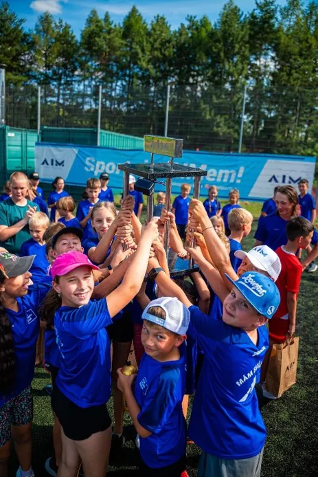 Skupina dětí v modrých tričkách drží společně trofej nad hlavami během venkovní sportovní akce.