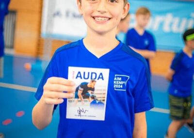 Chlapec v modrém tričku drží podepsanou kartu s obrázkem sportovce během akce v tělocvičně.