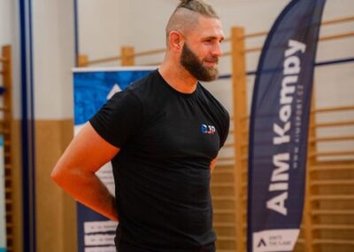 Jiří Procházka, známý MMA bojovník, stojí v tělocvičně během akce, oblečený v černém tričku a šedých kraťasech, s vlasy svázanými do drdolu a rukama za zády.