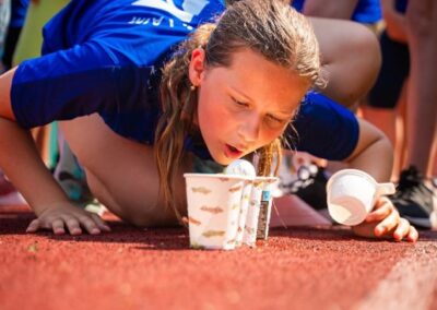 Dívka v modrém tričku se účastní venkovní soutěže, při které zvedá kelímek ústy na červené dráze, zatímco je podepřena rukou.